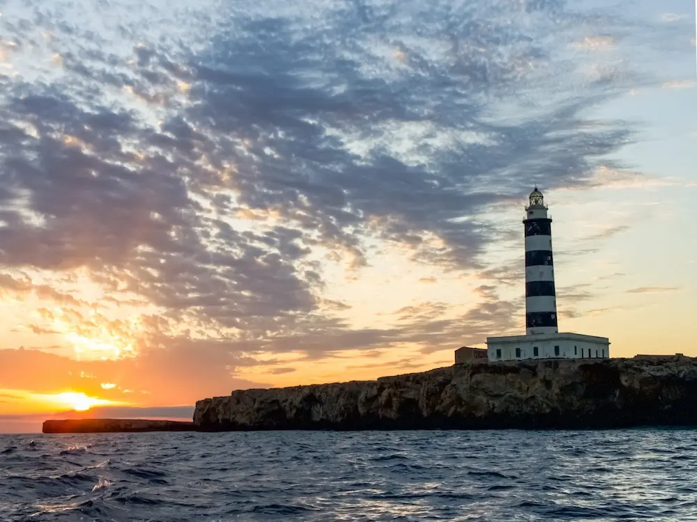 Isla del Aire lighthouse at sunset seen from the sea, with the sun setting over Menorca
