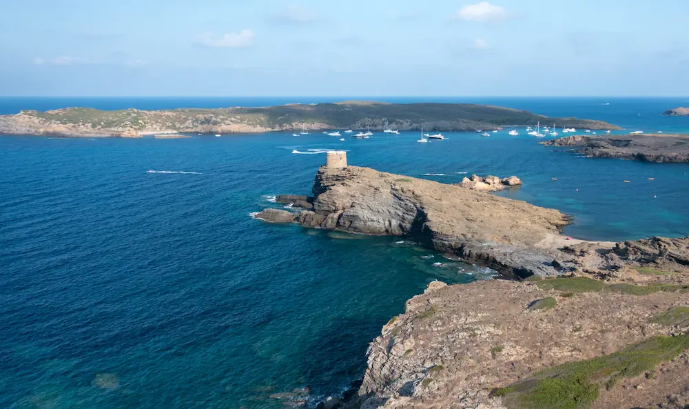 La Illa d'en Colom y una torre de defensa vistas desde la costa del parque natural de s'Albufera des Grau, con veleros fondeados