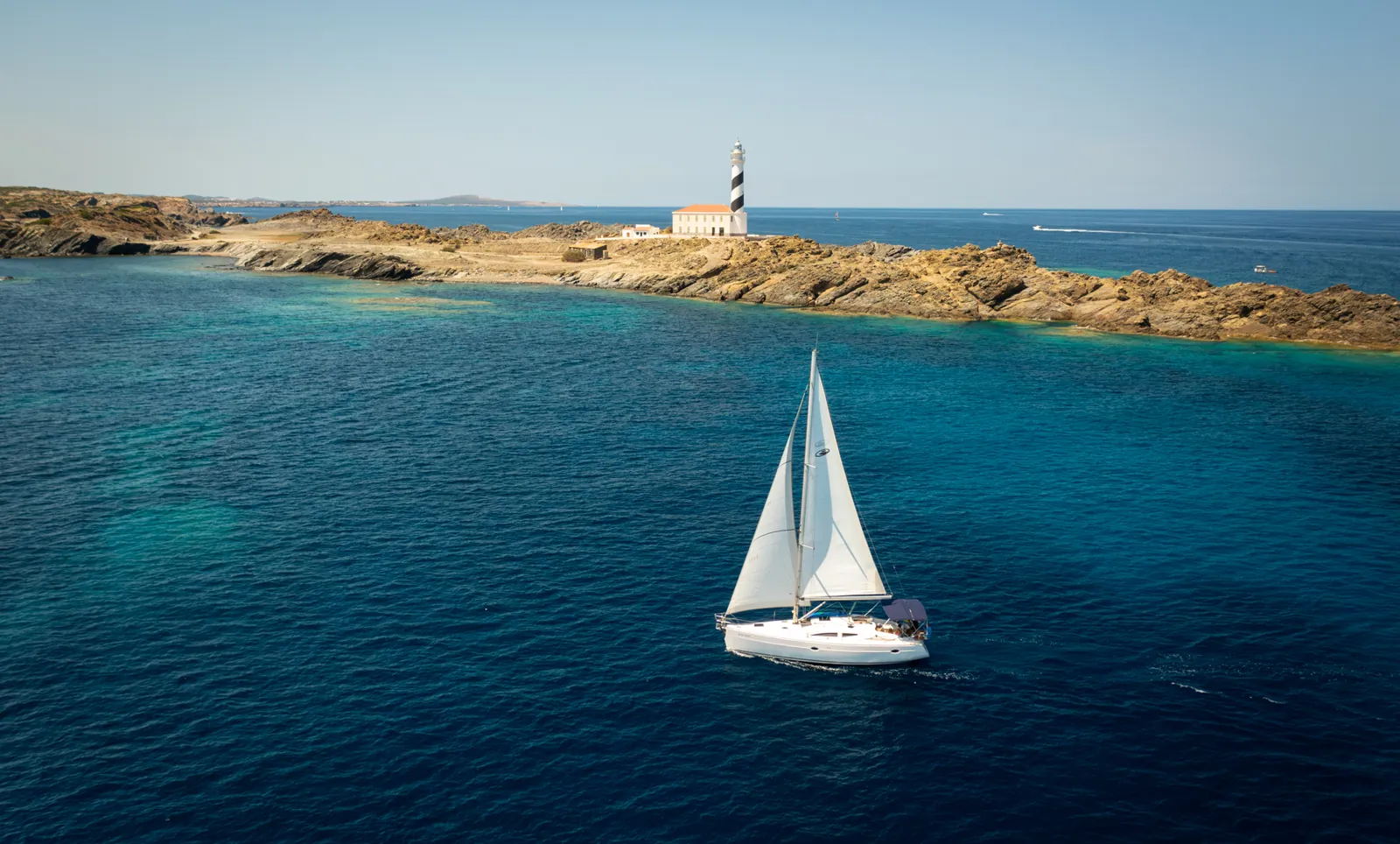 Sailboat sailing past the Favàritx lighthouse, over the black slate cliffs of northern Menorca